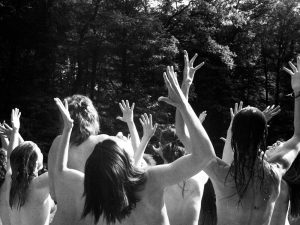 photo noir et blanc avec groupe de femme de dos torse nu les bras en l'air face à une fôret